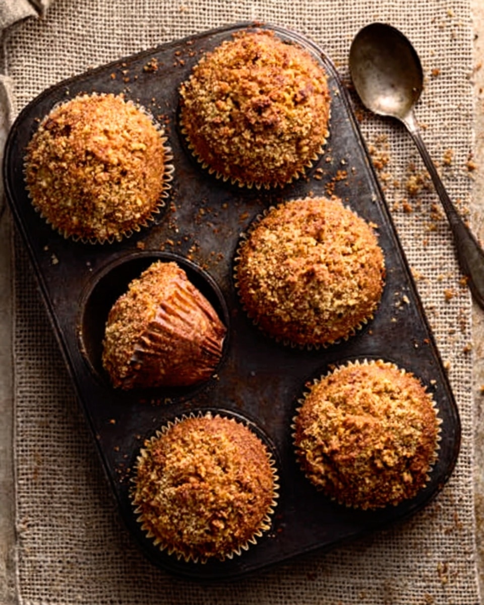 A dark metal muffin tray holds six muffins, each topped with a coarse, crumbly, golden-brown crust. The muffins rise high with some paper liners visible on the bottom left muffin, showing a subtle brownish pattern. The tray sits on a woven textured cloth with neutral earth tones. To the right of the tray is an old spoon with a rounded bowl resting on the cloth. The whole scene has warm lighting that highlights the crumbly texture on top of the muffins. photo taken with an iphone --ar 4:5 --v 7