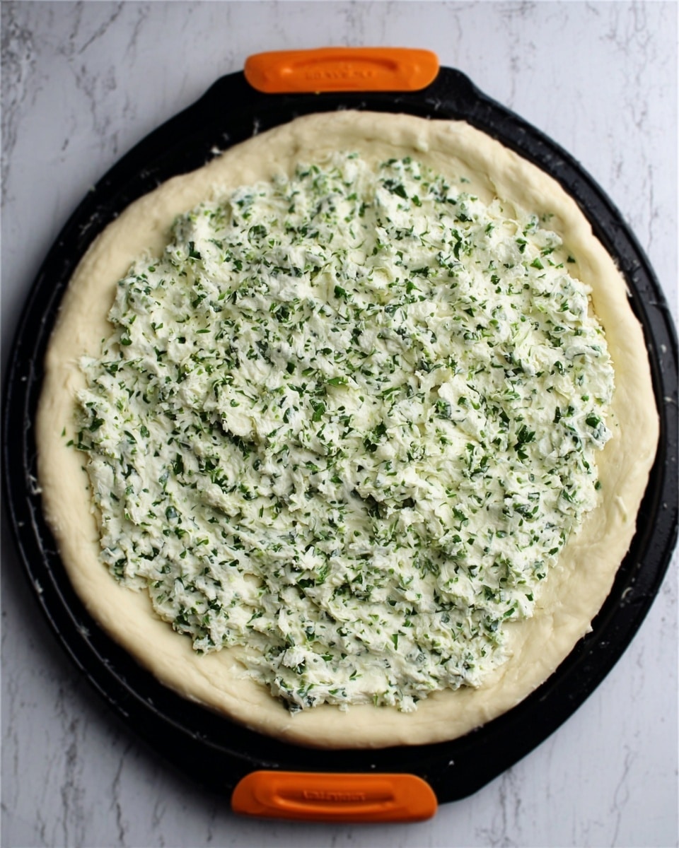 A round pizza dough spread evenly on a black baking tray with orange handles, topped with a thick, creamy white layer mixed with finely chopped green herbs, covering the entire surface almost to the edges. The dough has a slightly raised edge forming a crust around the topping. The tray rests on a white marbled surface. Photo taken with an iphone --ar 4:5 --v 7