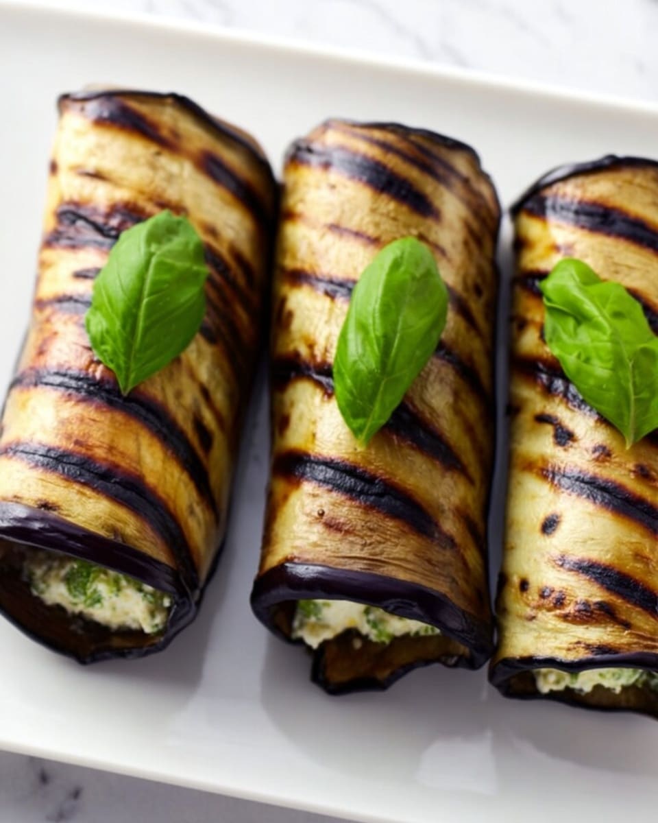 The image shows three rolled grilled eggplant slices arranged in a row on a white plate, each having dark grill marks that create horizontal stripes on a shiny, deep purple surface. Inside each roll, a light green creamy filling is slightly visible at the open ends. On top of the middle roll, there are three small bright green basil leaves placed as garnish. The background is a white marbled surface, adding a clean and bright look. photo taken with an iphone --ar 4:5 --v 7