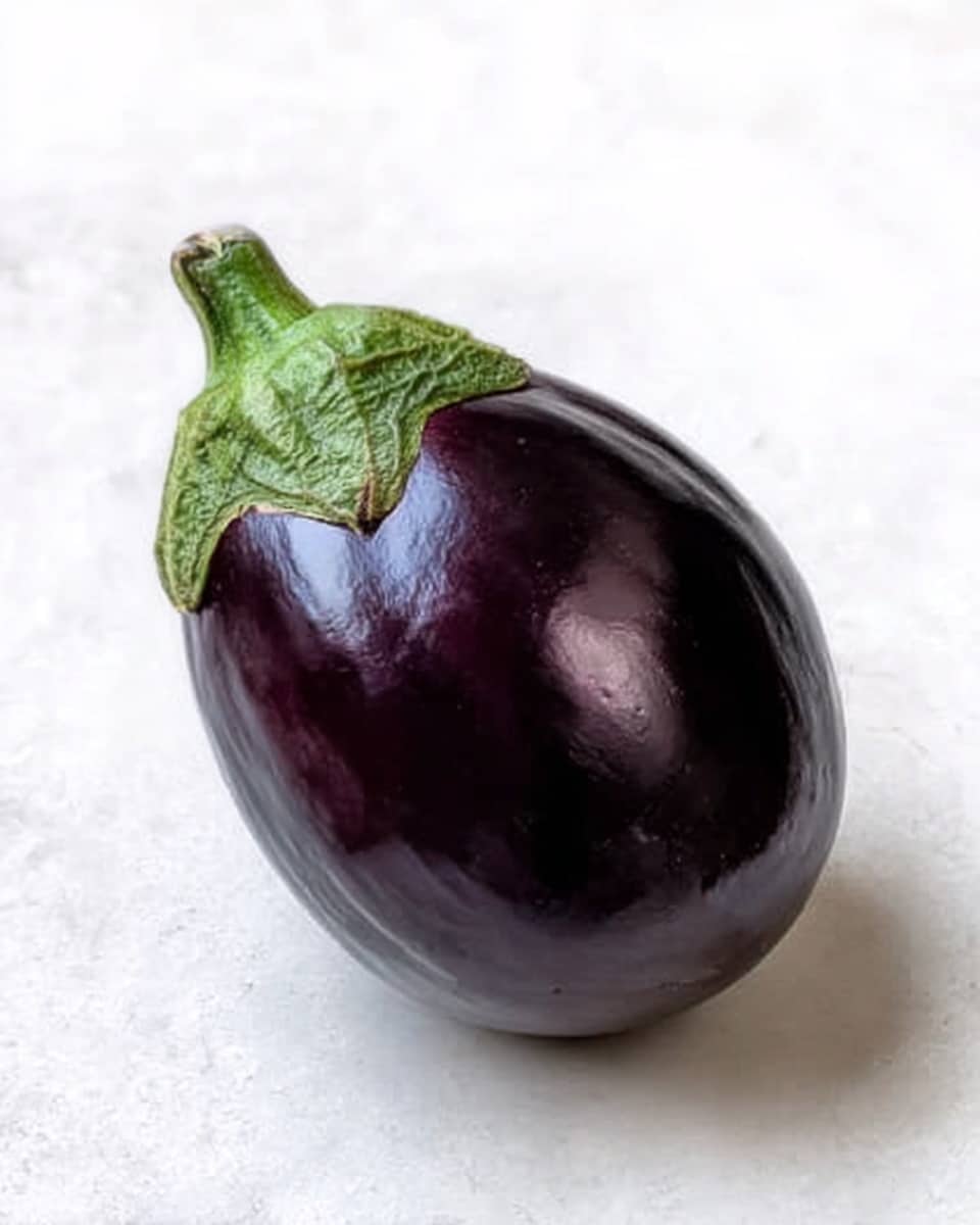 The image shows a single whole eggplant with shiny, smooth deep purple skin and a green calyx on top. The eggplant has an oval shape with a slightly tapered bottom. It is placed on a clean white marbled surface. The lighting highlights the glossy texture of the skin, making it look fresh and ripe. Photo taken with an iphone --ar 4:5 --v 7
