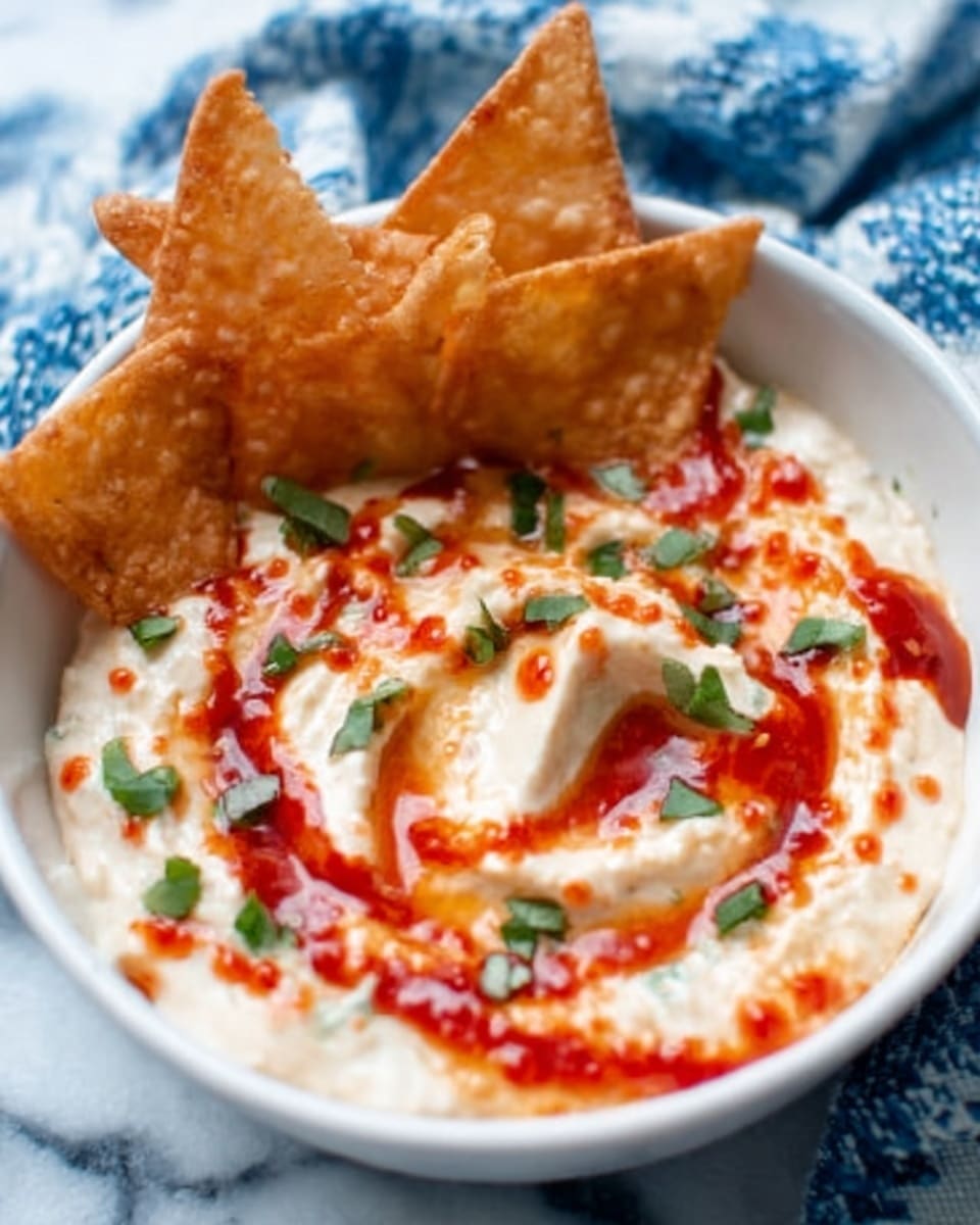 The image shows a white bowl filled with a creamy dip that has a swirled texture, appearing soft and smooth. The dip is topped with a drizzle of bright red sauce and small green herb pieces sprinkled on top. Around the edge of the bowl, several crispy triangular chips with a golden brown color lean against the side. The bowl is placed on a white marbled surface, with a blue and white cloth slightly visible in the background. photo taken with an iphone --ar 4:5 --v 7