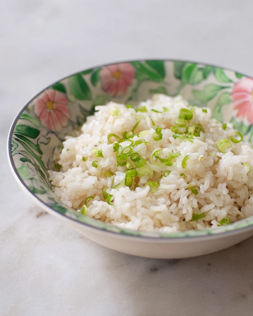 A bowl with a floral pattern holds a single layer of cooked white rice mixed with small chopped green onions scattered on top and within the rice, giving a fresh contrast of green and white. The bowl sits on a white marbled surface, and there is a slight soft shadow around the bowl edge. The texture of the rice looks fluffy and soft, with some grains sticking together. Photo taken with an iphone --ar 4:5 --v 7