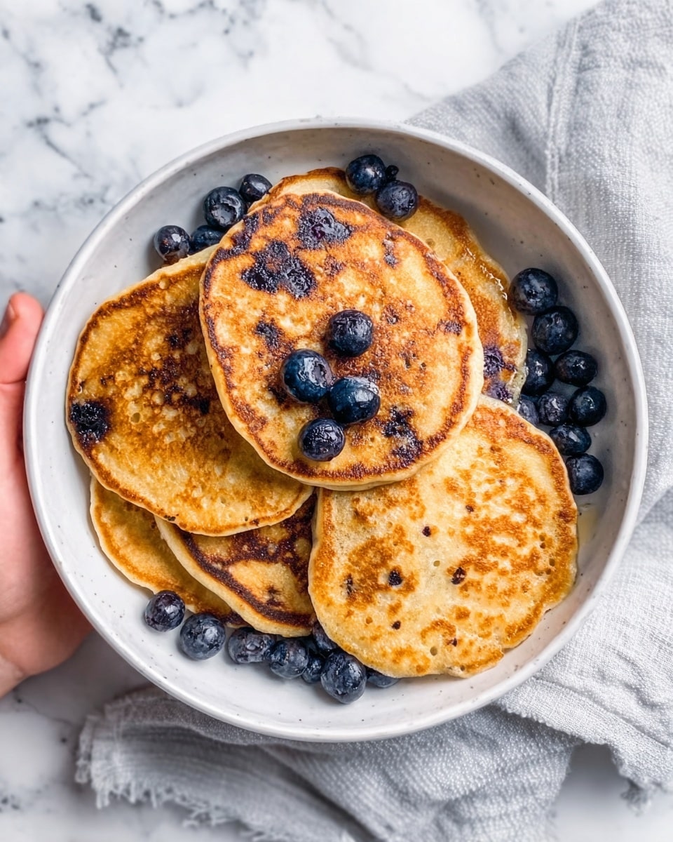 The image shows a white bowl filled with five golden-brown pancakes stacked loosely. Blueberries are placed both inside some pancakes, peeking through, and scattered around them in the bowl. The pancakes have a light, slightly uneven texture with some darker toasted spots. A woman's hand is holding the bowl from the side. The bowl sits on a white marbled surface with a light cloth partially visible nearby. Photo taken with an iphone --ar 4:5 --v 7
