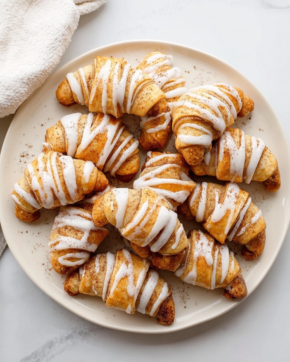 A white plate is filled with about twelve small, crescent-shaped pastries. Each pastry has a golden brown color with a slightly crispy looking texture. They are drizzled with thick, white icing in diagonal lines across the top. The pastries have a sprinkles of light cinnamon powder on both the dough and the icing. The plate sits on a white marbled surface with a white cloth partially visible in the top left corner. Photo taken with an iphone --ar 4:5 --v 7
