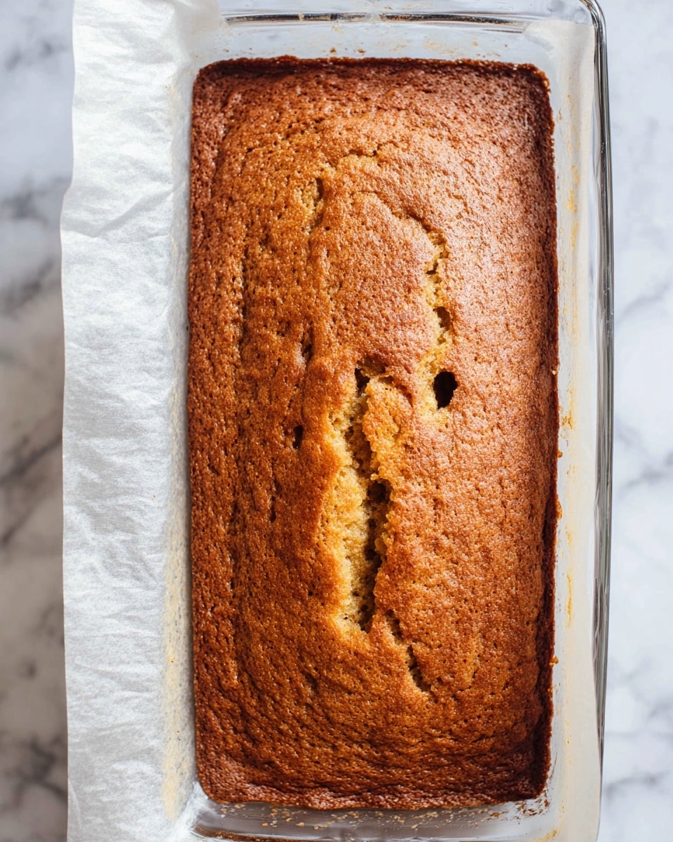 The image shows a close-up view of a freshly baked brown cake in a clear glass rectangular baking dish lined with parchment paper on the left side. The top of the cake has a rough, cracked texture with small holes and uneven areas, showing a golden-brown color with some darker spots. The cake fills the dish fully and the edges touch the sides. The background is a white marbled surface. photo taken with an iphone --ar 4:5 --v 7