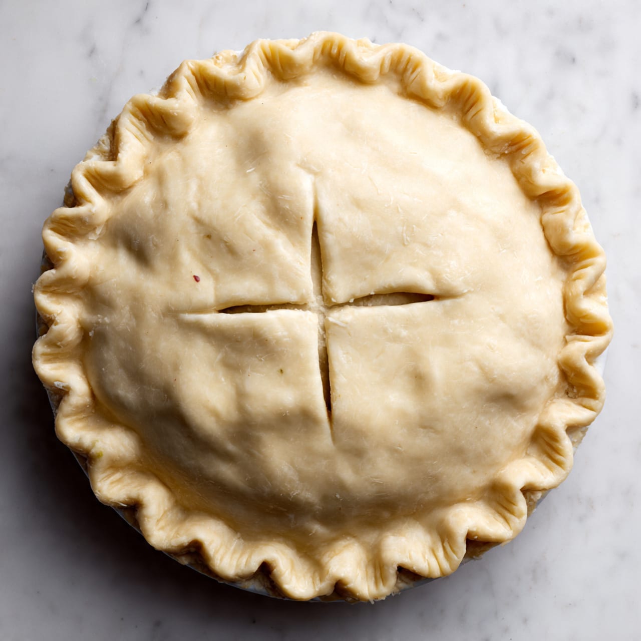 A whole round pie with a smooth, pale dough crust on top, edges pressed down with fork marks all around in an even pattern, and four short slits in the center forming an X shape to vent steam. The pie sits on a white marbled surface. The crust looks soft and unbaked, covering the filling completely and without any decorations or toppings. Photo taken with an iphone --ar 4:5 --v 7