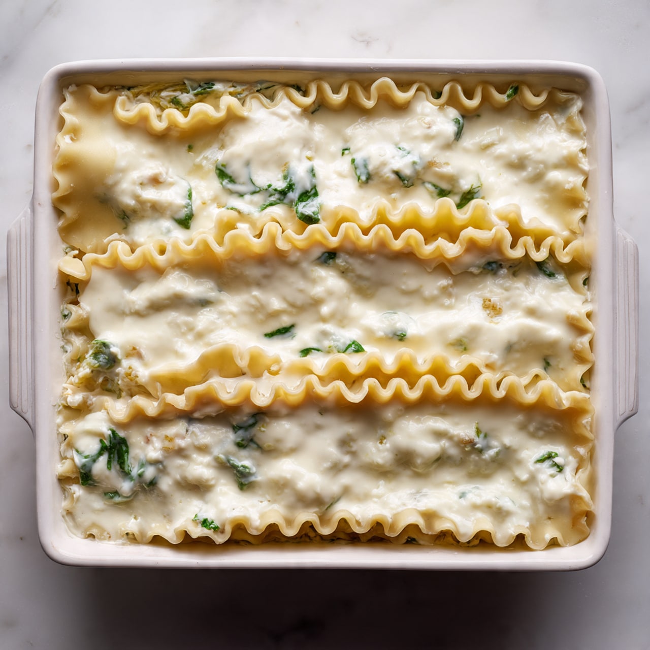 The image shows a white rectangular baking dish filled with three wide layers of uncooked lasagna sheets topped with a thick, smooth, creamy white sauce bubbling slightly on the surface. Beneath the lasagna sheets, small hints of green spinach leaves peek out from the edges, suggesting a layer of vegetables inside. The edges of the lasagna sheets have a wavy, slightly golden texture, contrasting with the milky sauce covering them. The whole dish sits on a white marbled surface. photo taken with an iphone --ar 4:5 --v 7