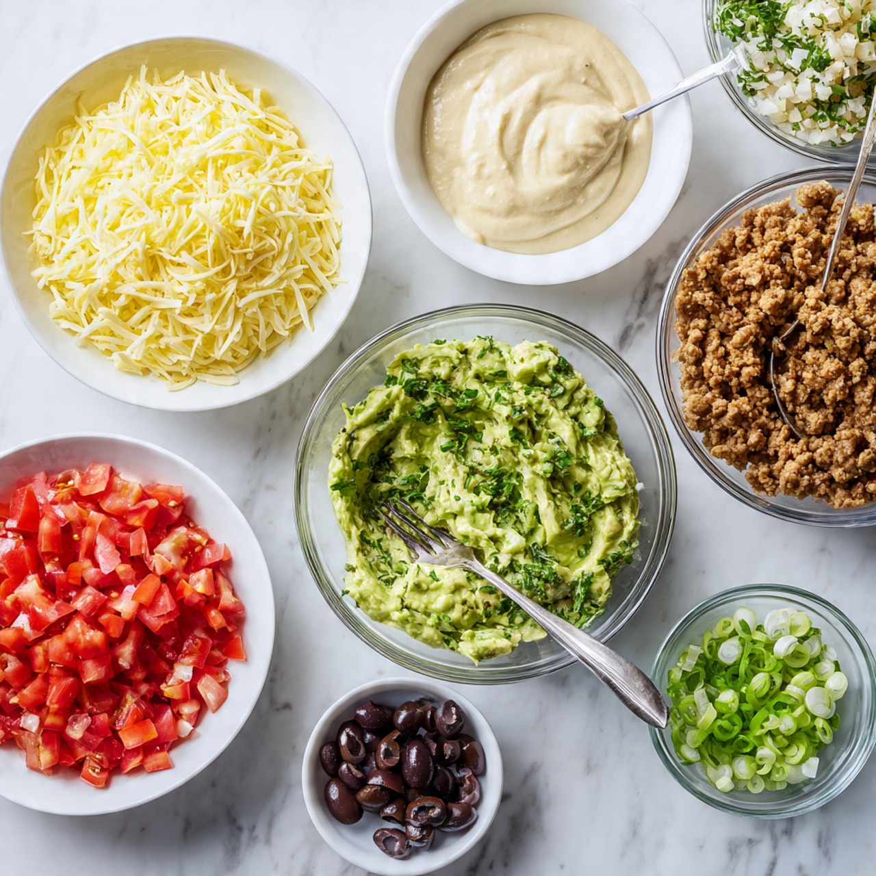 The image shows eight bowls arranged on a white marbled surface. At the center right is a large clear glass bowl, empty and shiny. To the top left is a white bowl filled with shredded yellow and white cheese. Below it is a large white bowl with mashed green avocado mixed with chopped herbs and a fork resting inside. To the top middle is a white bowl with creamy light beige sauce and a spoon standing in it. Next to it on the top right is a white bowl holding crumbled cooked brown protein with a spoon. Below the avocado bowl on the left is a white bowl filled with finely chopped red tomatoes. Between the tomato and the big bowl is a small white bowl with finely chopped dark brown olives. Below that is another small white bowl filled with sliced green onions. All items are bright and colorful on a clean white marbled background. photo taken with an iphone --ar 4:5 --v 7