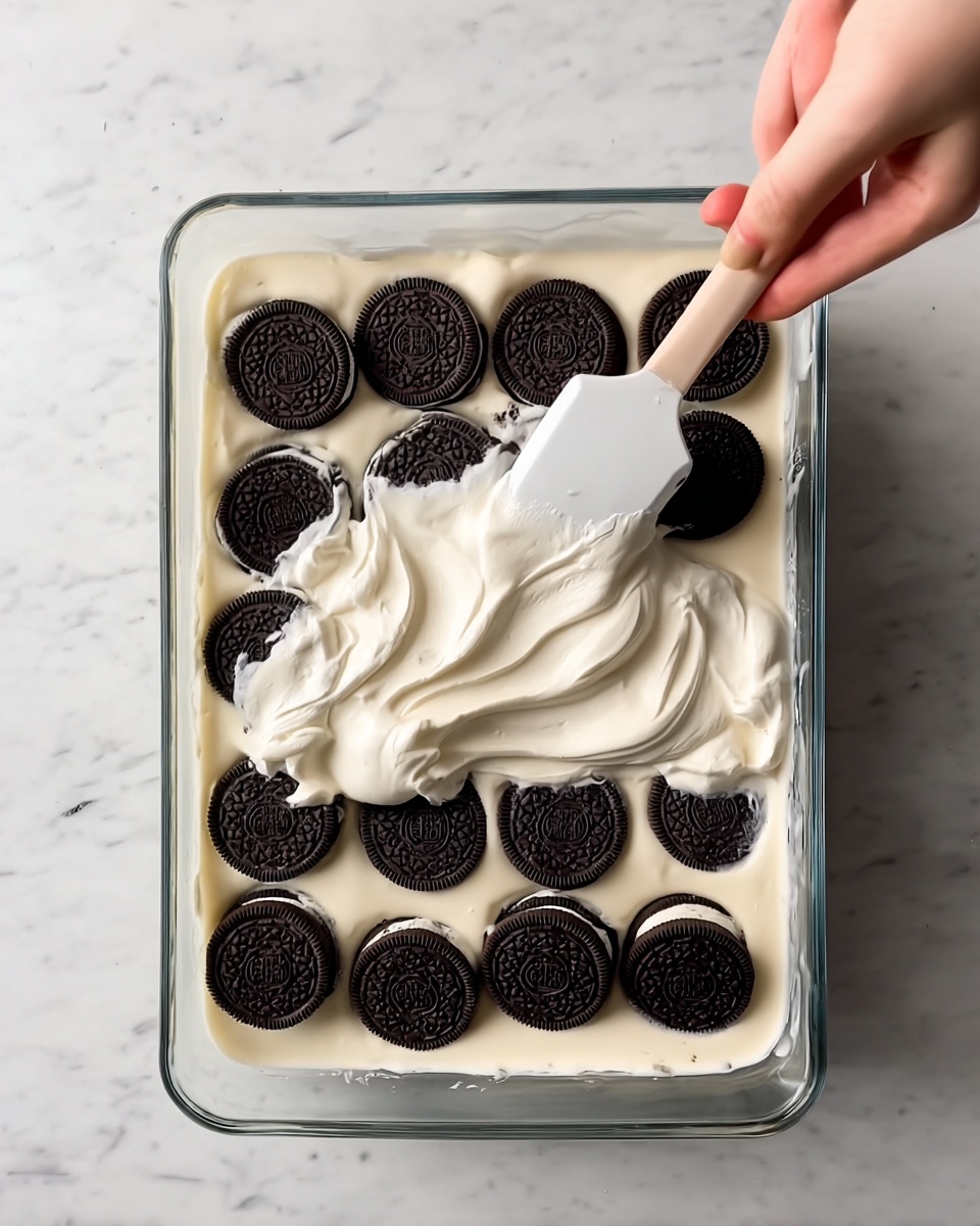 Oreo Dump Cake Recipe 4 A clear glass rectangular dish contains one layer of round dark chocolate sandwich cookies soaked in light beige creamy liquid, evenly spread at the bottom. A woman's hand holding a white spatula spreads a thick, smooth, white cream over the cookies, creating a fluffy top layer with soft texture. The clear dish is placed on a white marbled surface, adding a clean, bright background to the scene. photo taken with an iphone --ar 4:5 --v 7