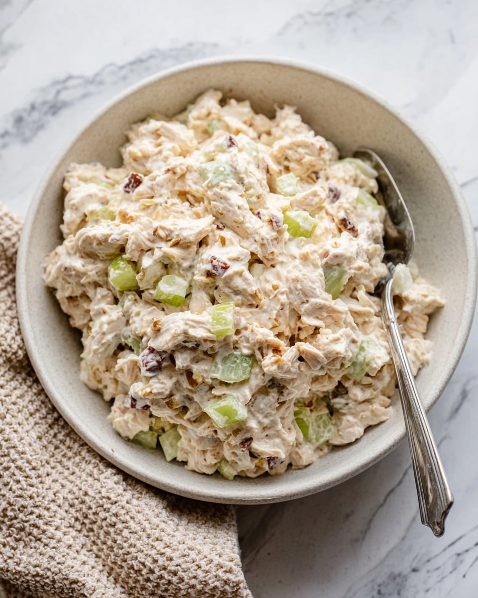The image shows a white bowl filled with a creamy chicken salad. The salad has small pieces of chicken mixed with finely chopped celery and small bits of something light brown, possibly nuts or onions. The chicken salad is soft and slightly chunky, giving a textured look. A silver fork rests inside the bowl on the right side. The bowl is placed on a white marbled surface, and to the left of the bowl, a beige textured cloth is partially visible. The colors in the salad are mainly off-white with green bits and light brown specks, creating a fresh and simple appearance. Photo taken with an iphone --ar 4:5 --v 7