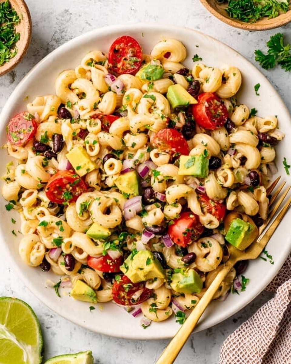 A white plate filled with a pasta salad that has three main layers: small curved pasta pieces in a light beige color, scattered chunks of bright red cherry tomatoes cut into halves, and larger pieces of light green avocado. There are also small black beans and bits of red onion mixed evenly throughout. The salad is garnished with fresh green herbs, adding a touch of color contrast. A gold fork rests inside the plate near the bottom right side. The background is a white marbled texture with some herbs and a halved lime partly visible at the bottom edge. Photo taken with an iphone --ar 4:5 --v 7