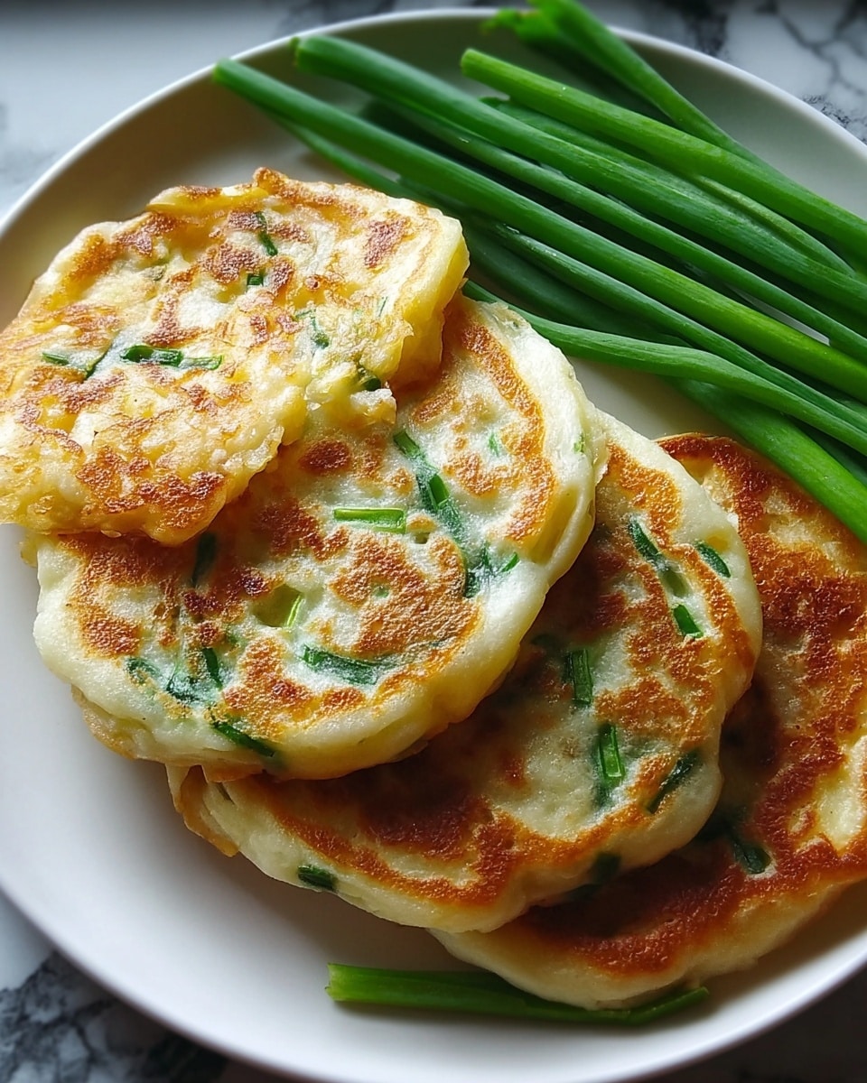 A white plate holds three golden brown, round pancakes stacked slightly overlapping each other. Each pancake has a crispy, bubbly texture with green onion pieces visible inside the soft dough. Next to the pancakes, several fresh green onion stalks lay parallel, adding a fresh green contrast. The plate rests on a white marbled surface. photo taken with an iphone --ar 4:5 --v 7
