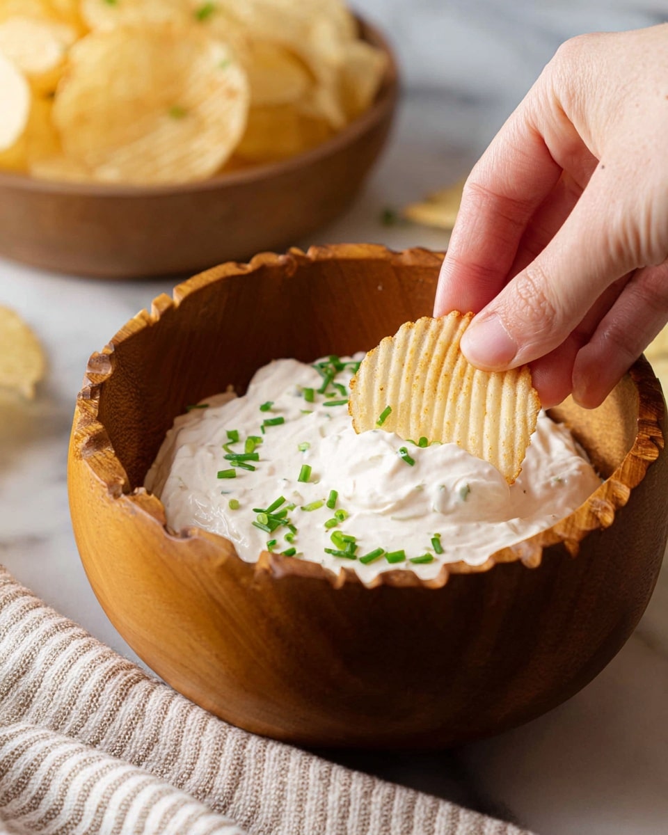 A woman's hand is dipping a ridged, light yellow potato chip halfway into a creamy white dip inside a large wooden bowl. The dip's surface is smooth with small green pieces scattered on top, likely chopped chives. The background shows a blurred bowl of similar potato chips, all set against a white marbled surface with a striped cloth partially visible near the bottom. Photo taken with an iphone --ar 4:5 --v 7