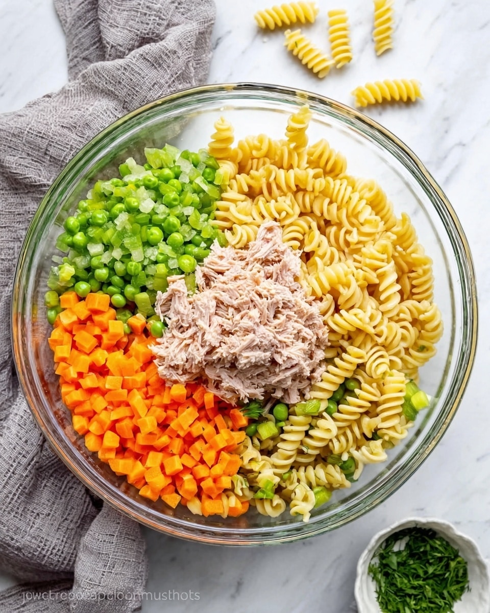A clear glass bowl sits on a white marbled surface, holding a colorful pasta salad with five main layers. Starting from the top right, there is a pile of pale yellow spiral pasta, next on the top left are small green chopped celery pieces, on the bottom left are bright orange chopped carrots, in the middle is a heap of light brown shredded tuna, and on the bottom right are vibrant green peas. A soft gray cloth is gently draped in the background, and a small white bowl with green herbs is visible at the bottom right corner outside the glass bowl. Photo taken with an iphone --ar 4:5 --v 7