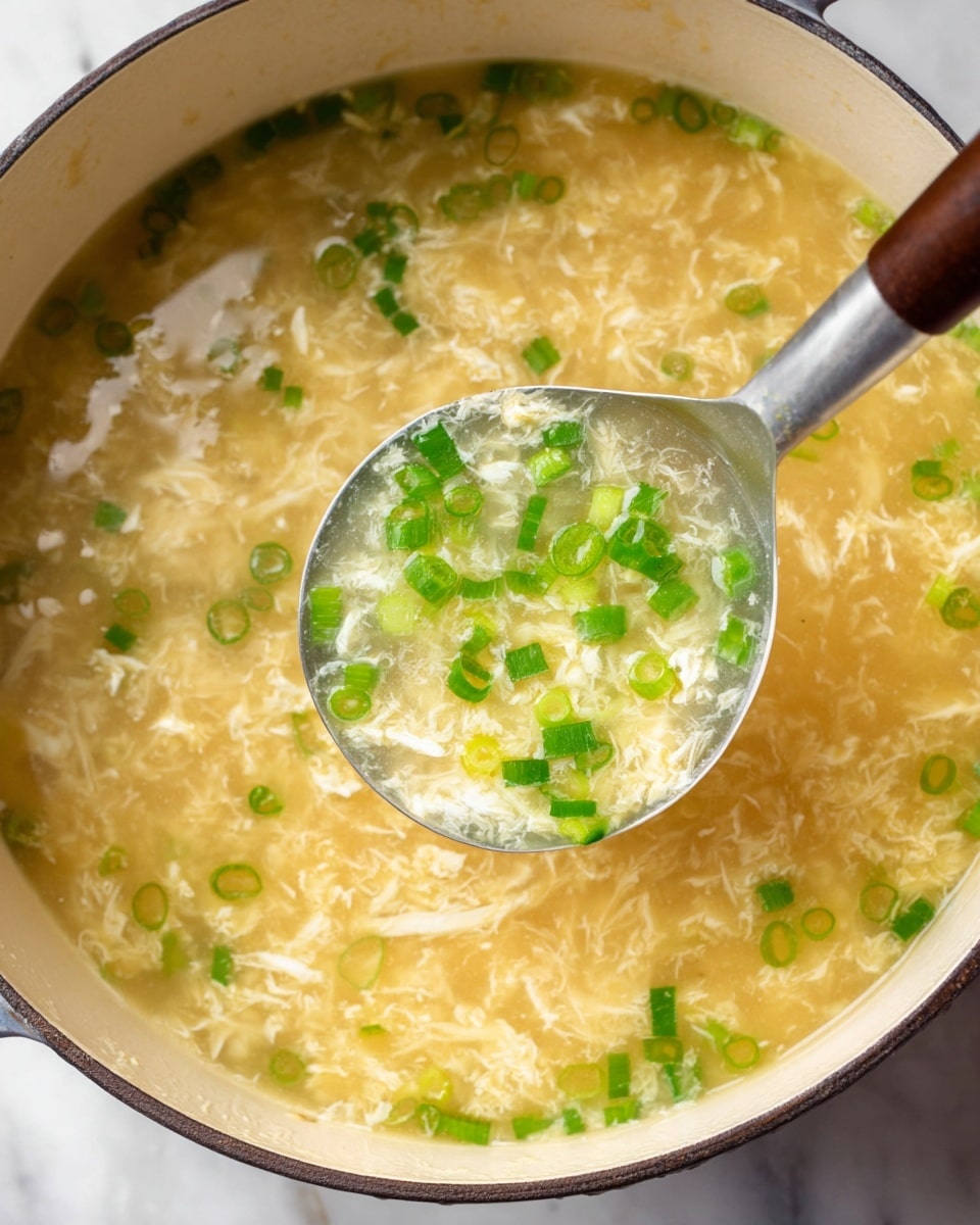 The image shows a pot filled with a light yellow egg drop soup with a slightly cloudy texture. Floating in the soup are finely chopped bright green spring onions, scattered evenly across the surface and inside the ladle. The ladle, made of silver metal with a brown handle, is held above the soup, showing the clear broth with white tangled egg strands and bits of green onions. The pot is white with a dark rim, sitting on a white marbled surface. Photo taken with an iphone --ar 4:5 --v 7