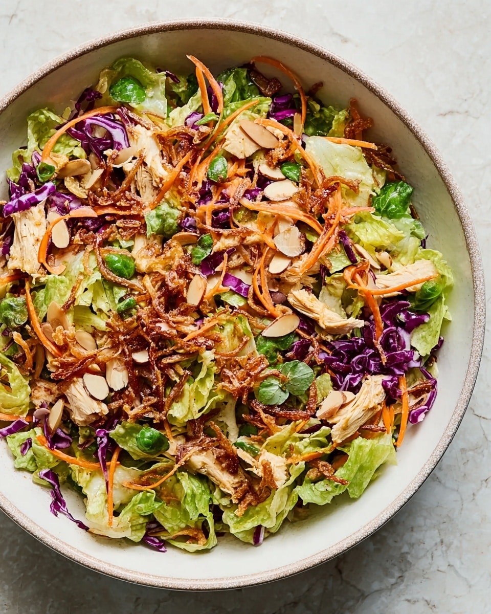 A bowl filled with a mixed salad showing layers of light green lettuce and purple cabbage with thin orange carrot strips scattered on top. There are pieces of light-colored chicken and toasted almond slices mixed throughout. Crispy fried onions add a golden brown touch, with bits of fresh green herbs sprinkled across the salad. The bowl is white with a subtle surrounding rim, placed on a white marbled surface. photo taken with an iphone --ar 4:5 --v 7