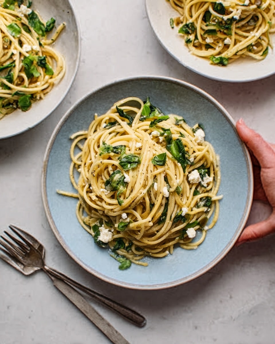 The image shows three white bowls on a white marbled surface, each filled with pasta. The pasta is light yellow with some dark green leaves mixed in. One bowl in the center is fully visible, holding spaghetti twisted into a neat mound with scattered pieces of what looks like feta cheese and small bits of herbs. A woman’s hand is reaching from the right side, gently holding this bowl. To the top left and top center behind this bowl, two more white bowls partially show similar pasta dishes with green leaves. A silver fork rests on the surface near the bottom left corner of the main bowl. The lighting is soft and natural, making the colors and textures very clear and fresh. photo taken with an iphone --ar 4:5 --v 7