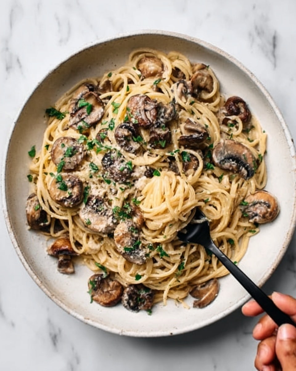 A round green pot with pale inside holds a serving of creamy beige spaghetti pasta as the base layer, spread evenly around the pot. On top, there is a thick layer of cooked dark brown mushroom slices clustered mostly on one half of the pasta, seasoned with small green herb bits scattered over both the mushrooms and pasta. A silver spoon with a red handle is placed to the right side inside the pot, resting on the pasta and mushrooms. The pot sits on a white marbled surface with a partial view of a white plate on the top right holding more of the same creamy pasta with mushrooms. Photo taken with an iphone --ar 4:5 --v 7