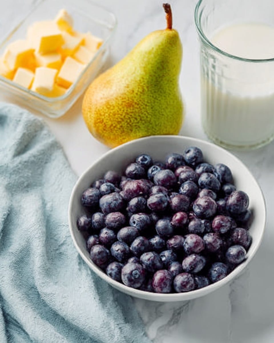 The image shows a white bowl filled with small round blueberries that have a deep purple color and a soft, slightly shiny texture. Next to the bowl on the white marbled surface is a pear with green and yellow skin and a small brown stem. To the left, there is a glass container holding pieces of light yellow butter with a creamy texture. A glass of milk with a white color is placed on the right side of the pear. In the background, a light blue cloth is softly folded. Photo taken with an iphone --ar 4:5 --v 7