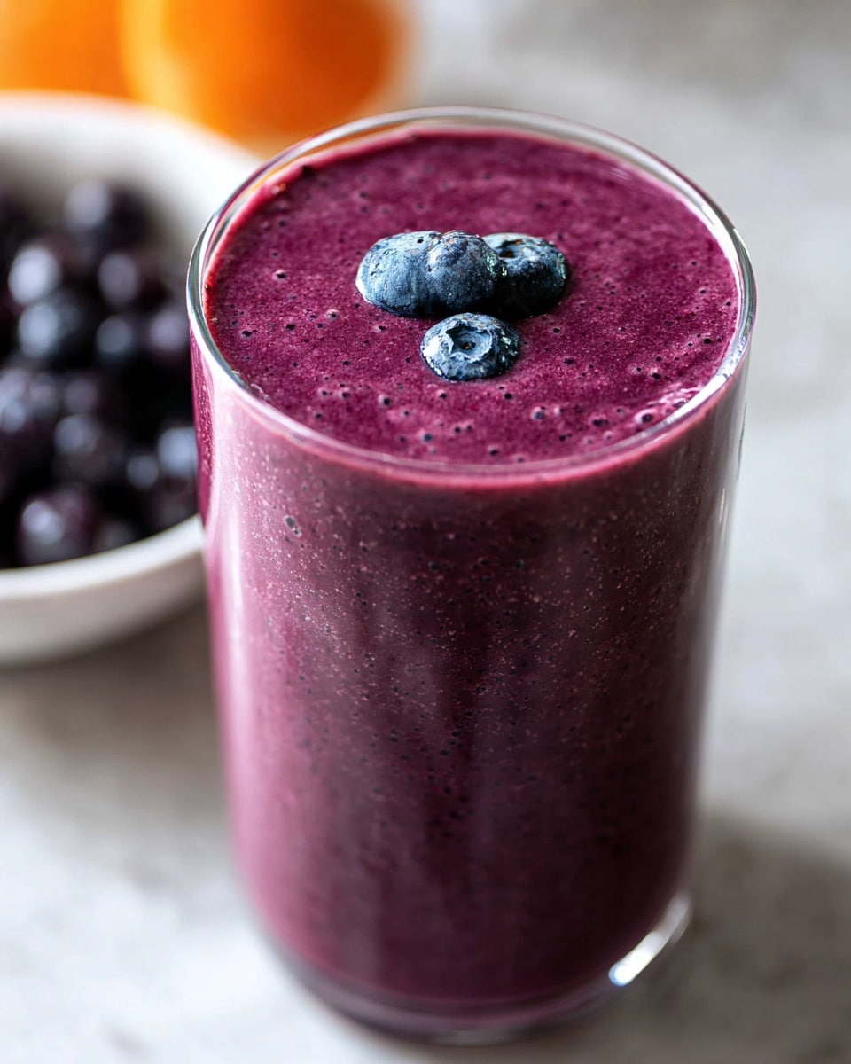 A tall clear glass is filled with a thick, deep purple smoothie that has a smooth texture with tiny bubbles on the surface. On top, there are three fresh blueberries, placed slightly off center. The glass sits on a white marbled surface, with a blurred white bowl filled with dark purple berries in the background. The lighting highlights the rich color and creamy texture of the smoothie. photo taken with an iphone --ar 4:5 --v 7