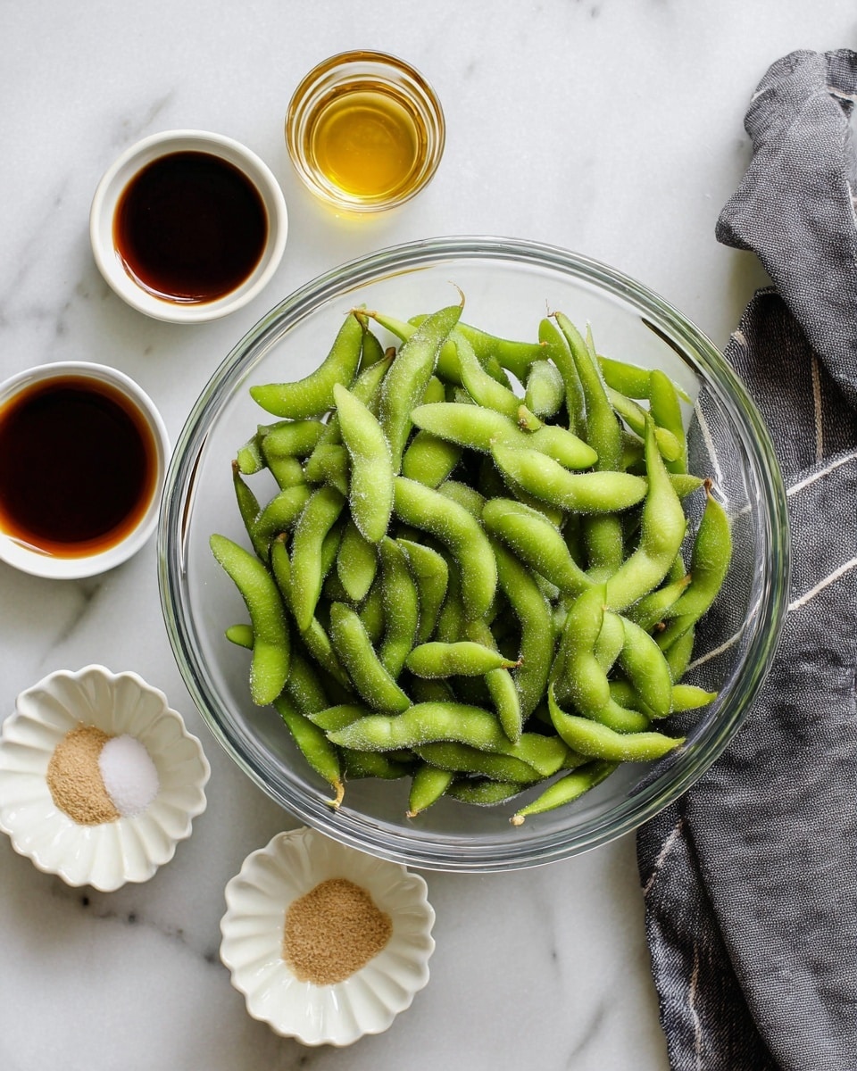 A clear glass bowl sits in the center filled with many bright green edamame pods, some pods showing slight frost texture. Around the bowl, there are six small white scalloped dishes arranged loosely, each holding different ingredients including dark brown soy sauce, light brown powder, coarse salt, and a golden liquid. A dark gray cloth with white stripes is partly visible on the white marbled surface in the upper right corner of the image. photo taken with an iphone --ar 4:5 --v 7
