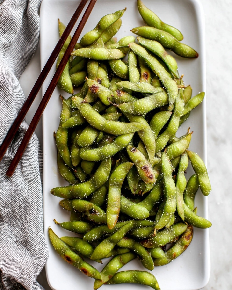 A white rectangular plate is filled with many green edamame pods that have a slightly wrinkled texture and some brown spots, sprinkled with coarse salt on top. The pods are arranged loosely, covering the entire plate surface. On the left side, a pair of dark brown chopsticks rest diagonally on the plate. The plate is set on a white marbled surface with a gray and white striped cloth partially visible on the left side. photo taken with an iphone --ar 4:5 --v 7