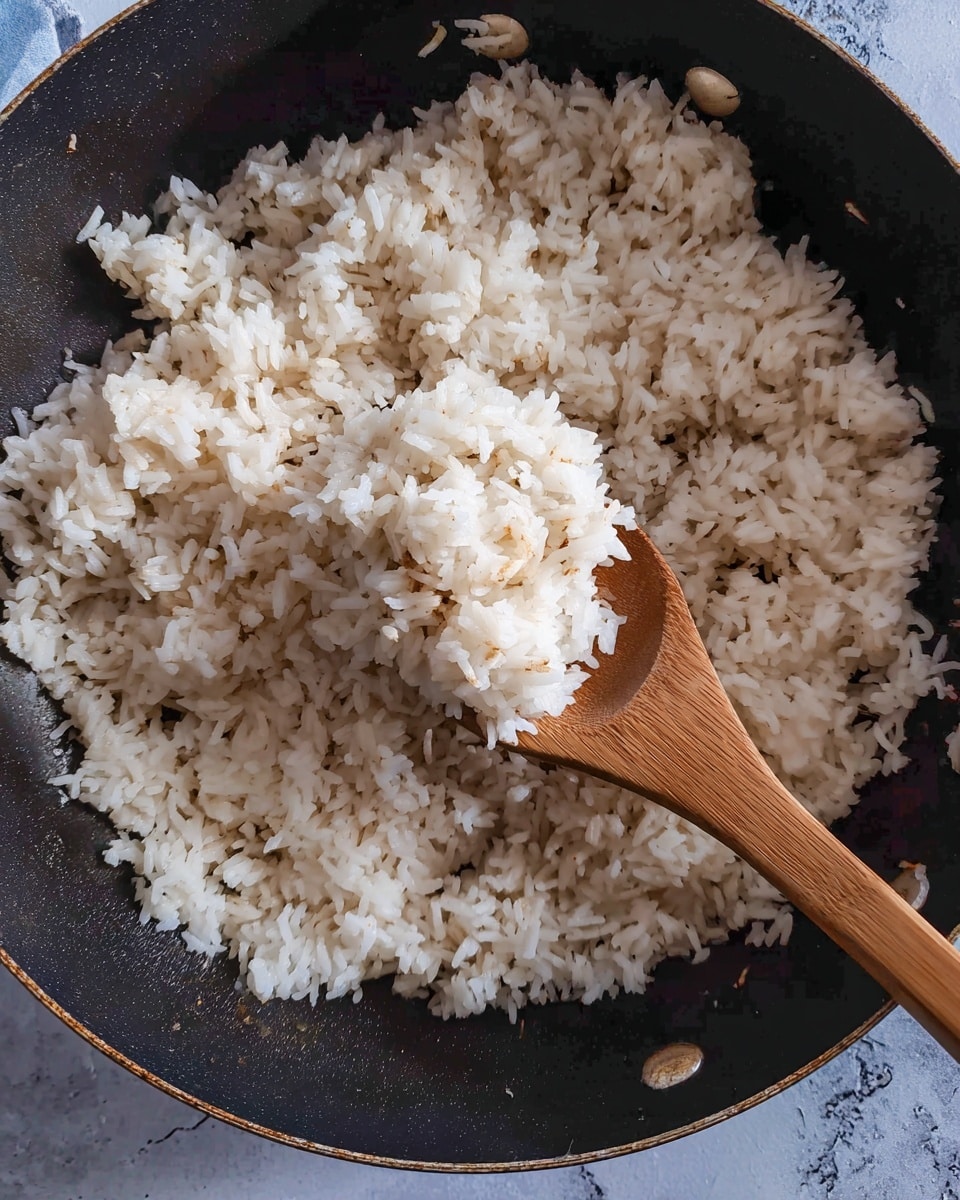 A close-up of a single layer of cooked white rice with small brown bits, all inside a large black pan. A wooden spoon is lifting some rice in the center, showing the rice grains' soft and slightly fluffy texture. The pan is on a flat surface styled with a white marbled texture. Photo taken with an iphone --ar 4:5 --v 7