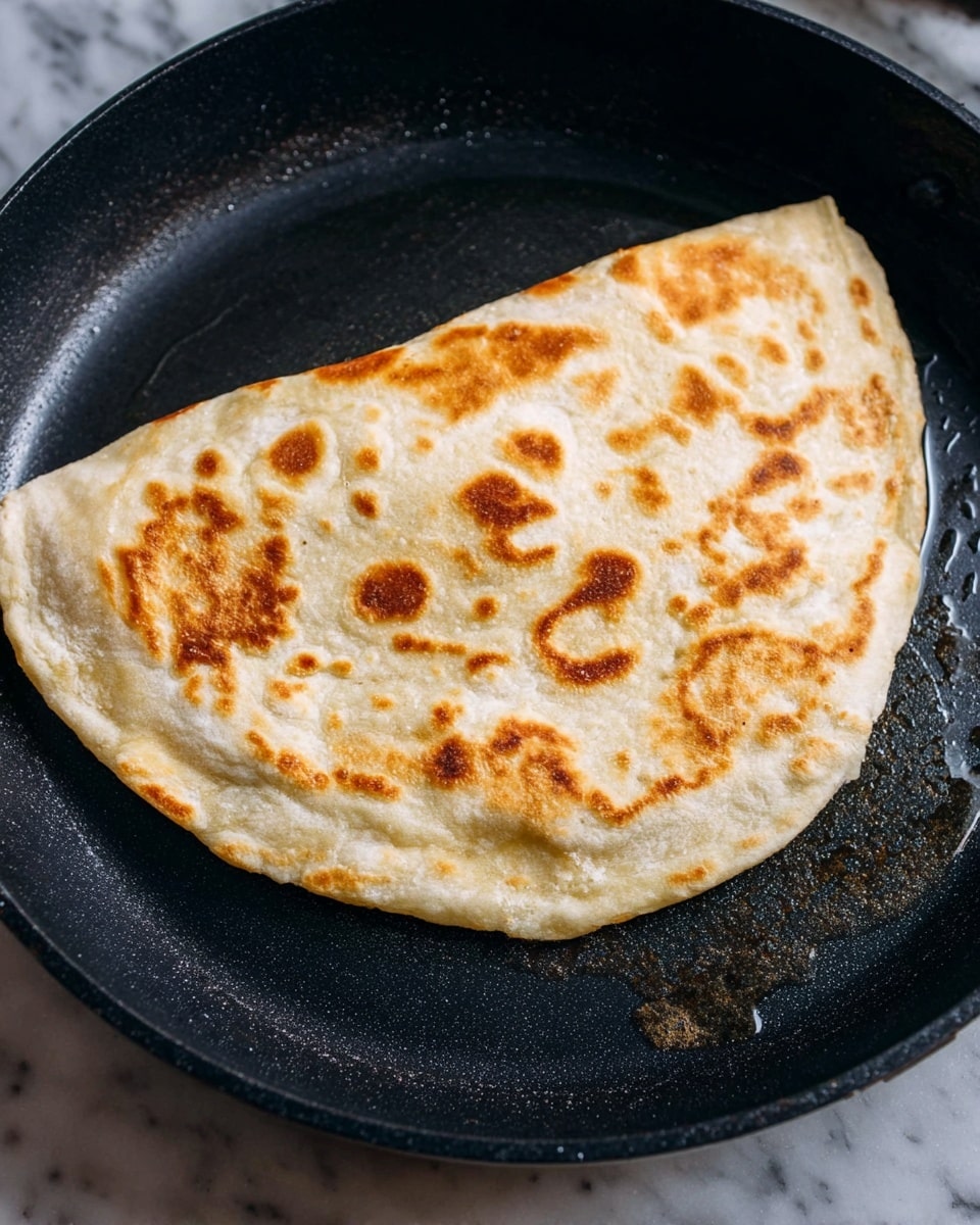 A single, half-folded flatbread with a light golden-brown toasted surface featuring uneven darker brown spots, sits in a black pan with a few drops of oil visible underneath. The flatbread’s edges are tucked and slightly thicker, with a soft, more doughy texture and a pale ivory color. The pan rests on a white marbled surface, adding contrast to the warm tones of the flatbread, photo taken with an iphone --ar 4:5 --v 7