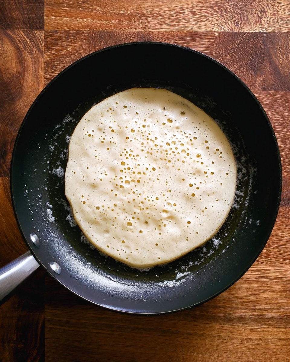 The image shows a round pancake cooking in a black frying pan placed on a wooden surface. The pancake batter is light beige with small air bubbles forming on the surface. The pan is slightly shiny with cooking oil spread evenly around the pancake. The wooden surface beneath has a rich, warm brown tone with a textured pattern. photo taken with an iphone --ar 4:5 --v 7