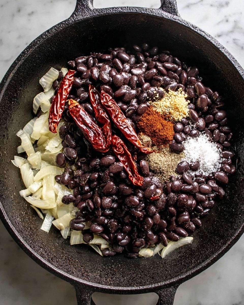 A black cast iron pan holds layers of ingredients starting with a base of pale, sliced onions spread evenly at the bottom. On top of this, there is a thick pile of dark, glossy black beans covering most of the center. Above the beans are a few wrinkled deep-red dried chili peppers placed near the top right side. Around the chili peppers, there are small piles of brown and light tan spices, along with a sprinkling of white salt, all resting on the black beans. The pan sits on a white marbled surface. photo taken with an iphone --ar 4:5 --v 7