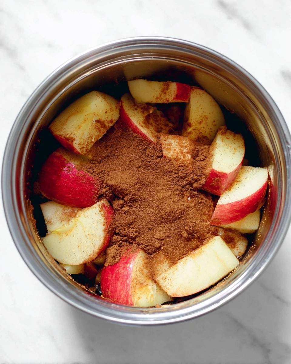 A metal container filled with cut apple pieces mostly showing their white flesh and red skin, topped with a generous layer of brown cinnamon powder. The apple pieces are partly covered in a dark liquid, giving a glossy texture, and the container sits on a white marbled surface. The cinnamon powder appears finely ground, creating a soft, crumbly texture over the fruit. photo taken with an iphone --ar 4:5 --v 7