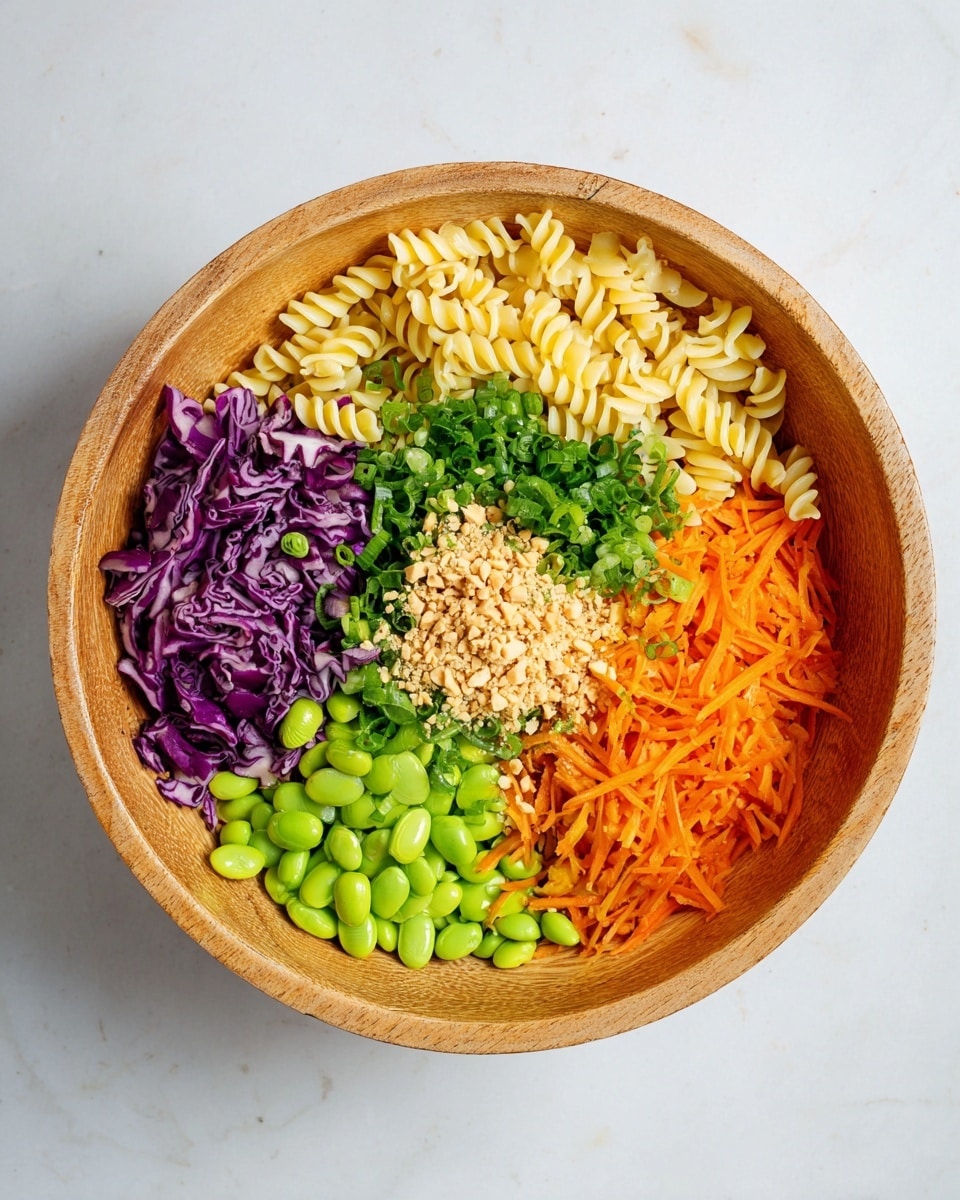 A large round wooden bowl holds six distinct layers of ingredients arranged side by side, filling the bowl evenly. On the top left, there is a portion of pale yellow spiral pasta with a smooth texture. Next to it, moving clockwise, is a bunch of finely chopped green herbs, bright and leafy. Adjacent to the herbs is a section of thinly sliced purple cabbage with a crisp texture. Below the cabbage, vibrant orange shredded carrots form a thick layer. To the left of the carrots, a pile of shiny green edamame beans adds a fresh look. In the center of the bowl, finely chopped light green onions are topped with a small mound of crushed light brown peanuts, creating a textured center. The bowl sits on a white marbled surface. Photo taken with an iphone --ar 4:5 --v 7