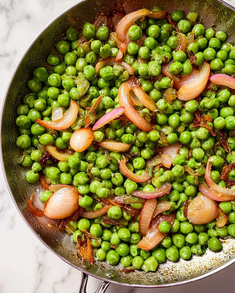 A pan filled with bright green peas mixed with caramelized light brown and pinkish onion slices scattered evenly across the dish, each onion piece showing a soft, slightly shiny texture. The peas look fresh and round, with a light glaze giving them a subtle shine. The pan has a brushed metal surface with some oil spots, creating a warm contrast to the peas and onions. The background is a white marbled texture. Photo taken with an iphone --ar 4:5 --v 7