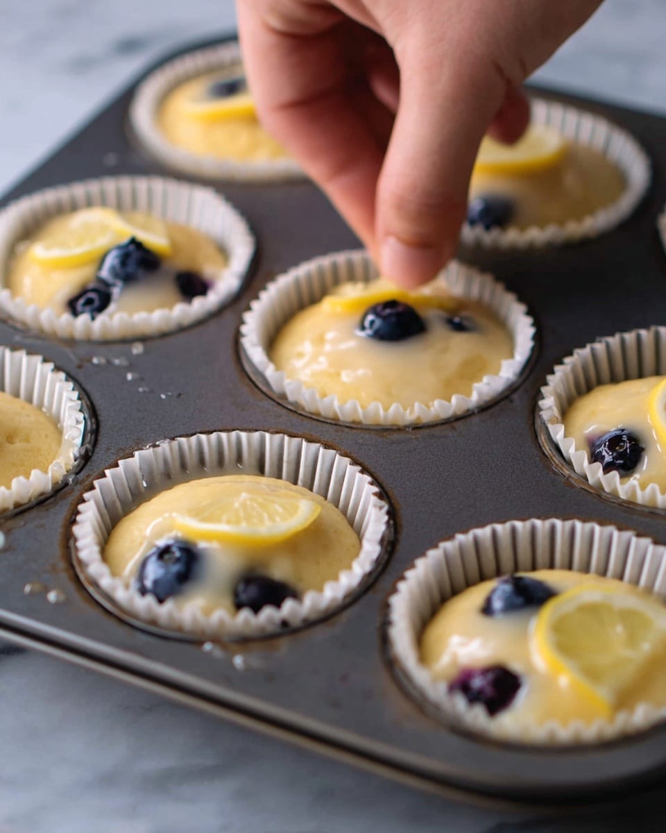 A close-up view of a dark metal muffin tray filled with light yellow batter inside white paper liners. Each liner holds a few dark blueberries and some have a thin slice of lemon on top. A woman's hand is gently placing a small amount of clear jelly or glaze on one of the muffins near the front left. The tray rests on a white marbled surface, and the batter has a smooth, slightly shiny texture. photo taken with an iphone --ar 4:5 --v 7