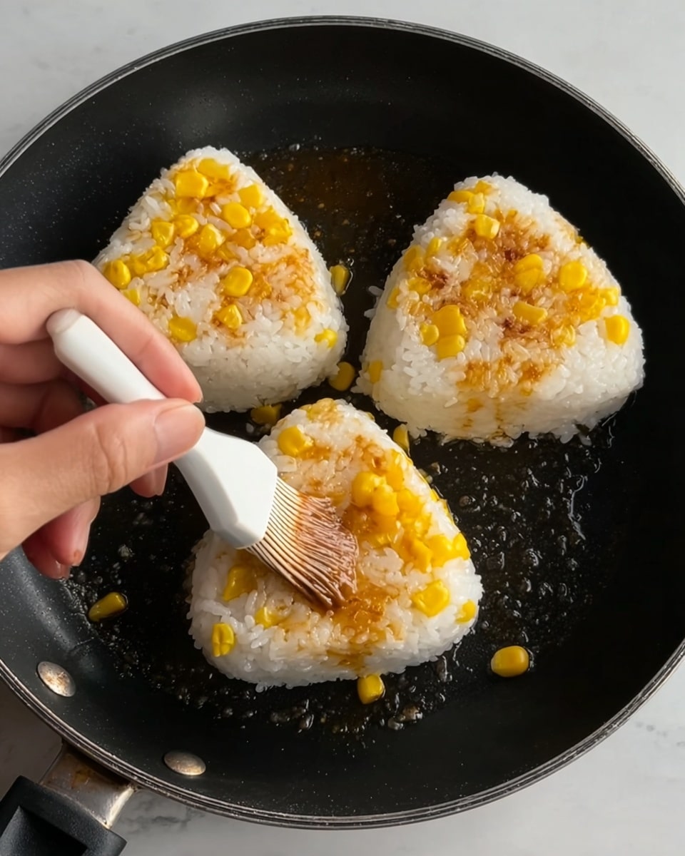 Three rice balls mixed with bright yellow corn kernels are in a black frying pan over a white marbled surface. Each rice ball is triangular with a slightly browned, crispy side facing up. A woman's hand is brushing a light brown sauce onto the closest rice ball using a white silicone brush. The rice is white and sticky with the corn pieces scattered evenly inside. Photo taken with an iphone --ar 4:5 --v 7