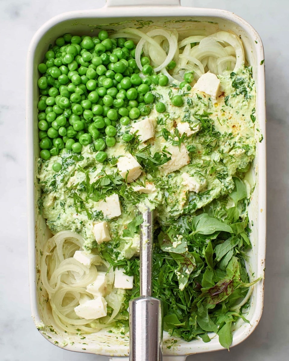 A white rectangular dish holds several layers of fresh ingredients ready to be blended with a metal hand blender. On the left side, there are bright green peas and translucent cooked onion slices scattered around. The center is covered with a light green creamy mixture mixed with bits of green herbs. Fresh dark green leafy herbs are placed on the bottom right, while small pieces of white cheese are unevenly spread near the top left. The dish sits on a white marbled surface. Photo taken with an iphone --ar 4:5 --v 7