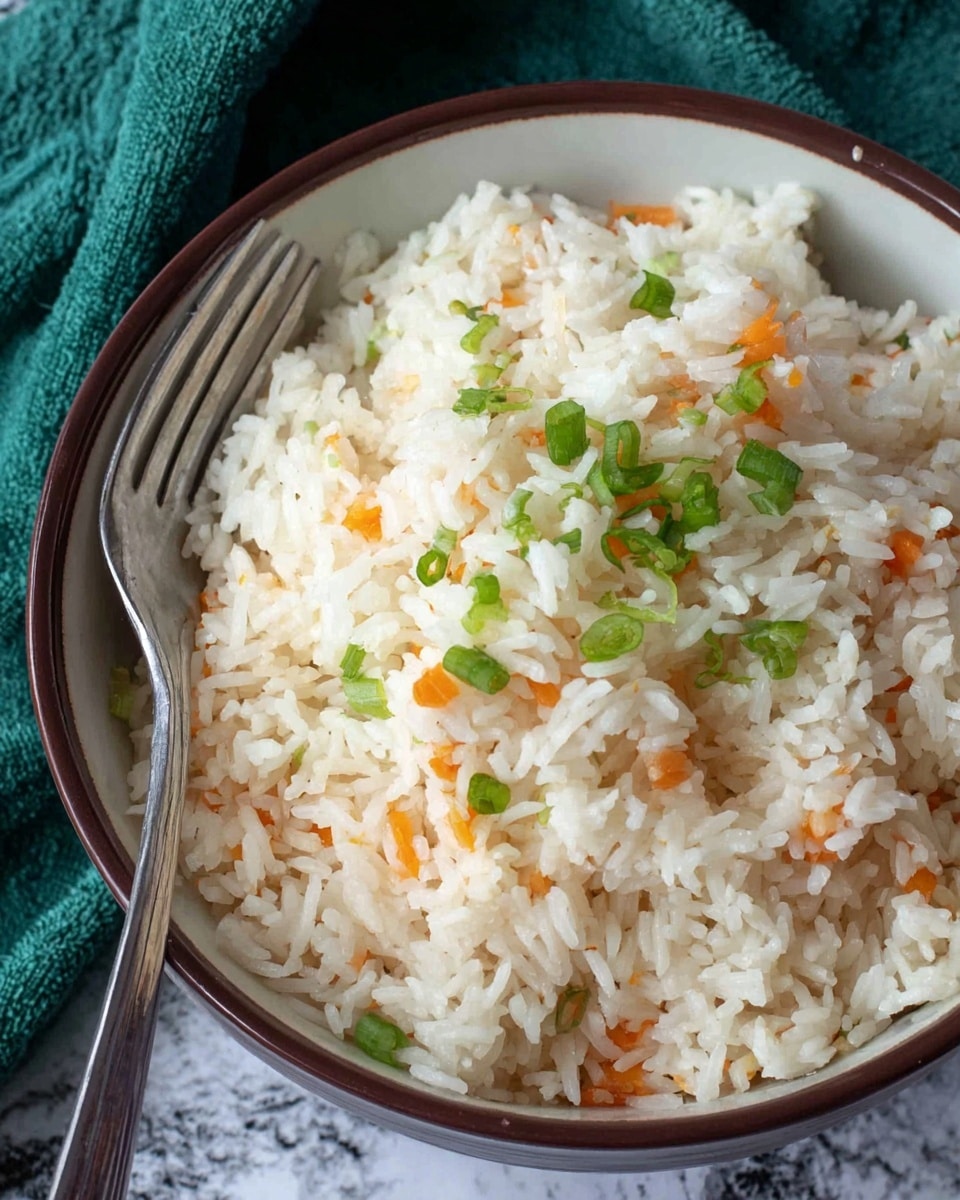 A close-up view of a bowl filled with white cooked rice mixed lightly with small bits of orange carrot and sprinkled with chopped green onions on top, all inside a white bowl with a brown rim. A silver fork is placed on the left side, partially inside the bowl. The background is a white marbled texture with a green cloth partially visible near the bowl photo taken with an iphone --ar 4:5 --v 7