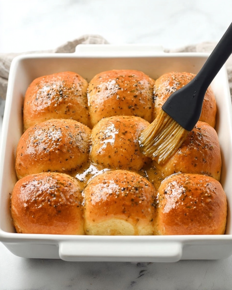 A white rectangular baking dish holds twelve soft, golden brown rolls arranged in three rows of four. The rolls are coated evenly with a shiny, thick, light brown sauce speckled with black pepper. A black brush with a black handle is brushing the sauce on the rolls from the lower left corner, resting on the white marbled surface beneath. The rolls have a smooth, glossy texture with light ridges on top. Photo taken with an iphone --ar 4:5 --v 7