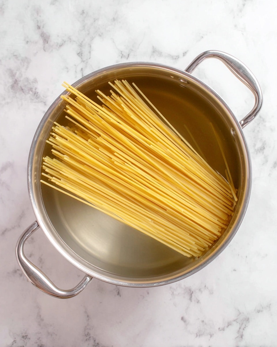 A large silver pot filled with clear water sits on a white marbled surface, with uncooked yellow spaghetti partially submerged inside. The spaghetti strands are straight and grouped closely together in the center, with their ends resting against the side of the pot. The pot has two silver handles on each side and reflects soft light on its smooth metal surface. Photo taken with an iphone --ar 4:5 --v 7