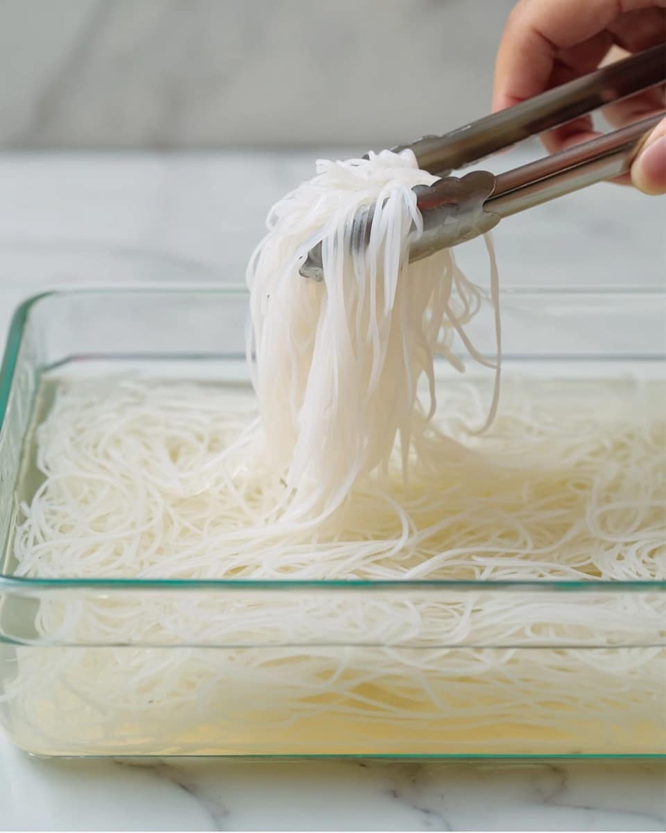 A close-up image shows a rectangular transparent glass container filled with clear water and white flat rice noodles soaking inside. A pair of metal tongs held by a woman's hand is lifting a bunch of the soft noodles out of the water, causing the noodles to clump together with a smooth and slightly wet texture. The background has a white marbled texture. photo taken with an iphone --ar 4:5 --v 7