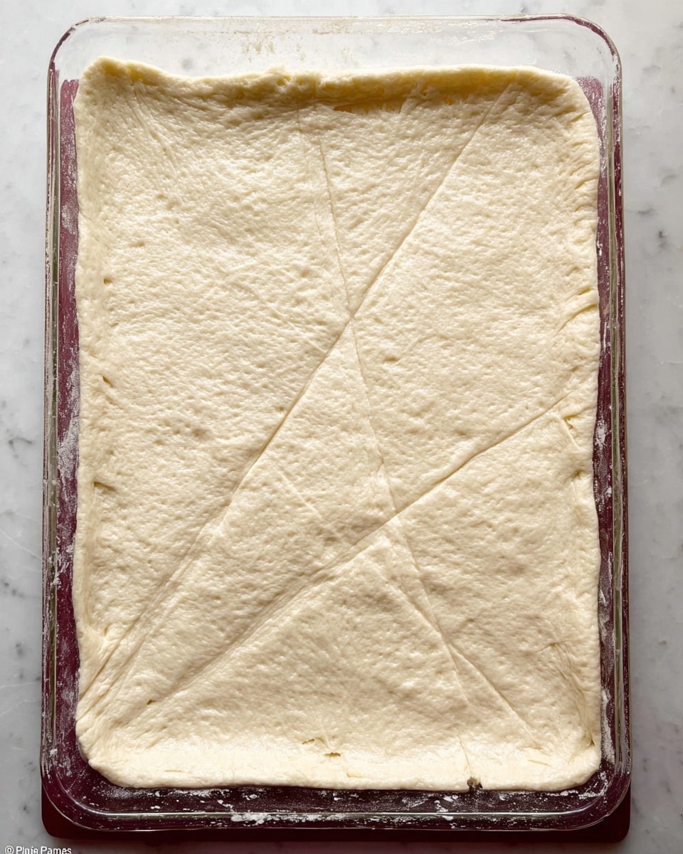 A clear glass rectangular baking dish holds a large, flat layer of pale, raw dough stretched to fill the dish almost edge to edge. The dough surface is smooth with slight visible creases and a few small air bubbles. The edges are lightly pressed against the dish, and there is a simple line of small holes forming a horizontal indentation near the middle, as well as faint diagonal lines crossing the dough. The dish is set on a white marbled surface. photo taken with an iphone --ar 4:5 --v 7