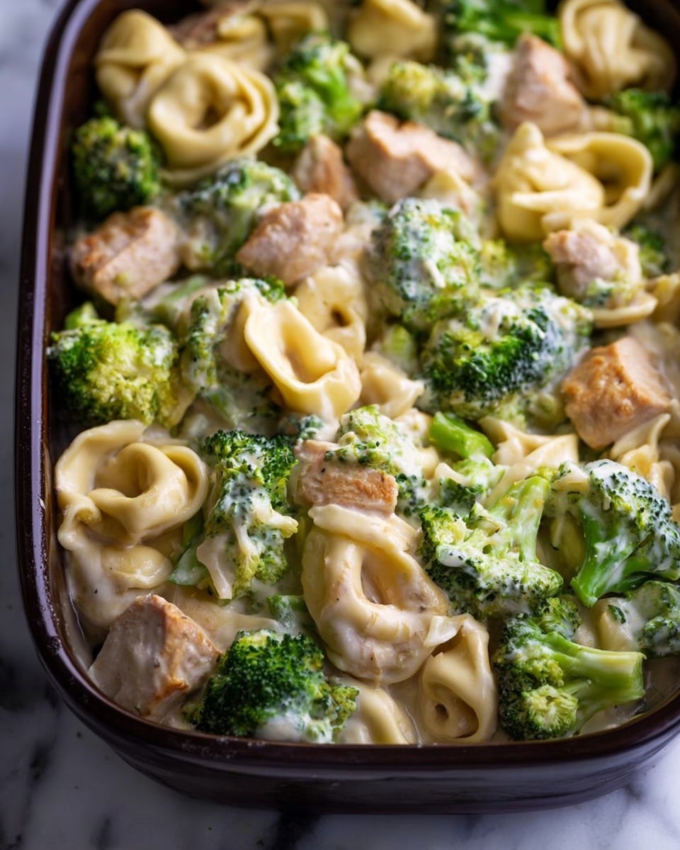 The image shows a large dark brown baking dish filled with a creamy pasta casserole. The dish has three main layers mixed together: small tortellini pasta in a pale yellow color, light beige pieces of cooked chicken evenly spread, and bright green broccoli florets scattered throughout. The creamy sauce coats all ingredients, giving a shiny and smooth texture over the pasta, chicken, and broccoli. The dish sits on a white marbled surface next to a wooden spoon on the right and a white plate with a silver fork on the upper right corner. photo taken with an iphone --ar 4:5 --v 7