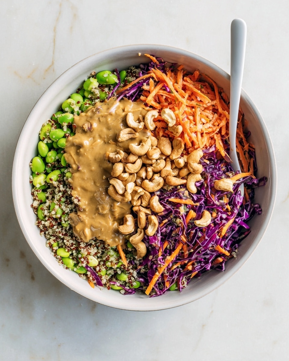 A white bowl holds a colorful salad with four main layers. The first layer is a mix of green edamame, orange shredded carrots, and purple cabbage strips spread evenly. The second layer features small, light beige quinoa grains scattered throughout. On one side, a cluster of whole roasted cashews with a slight brown tint is piled up. Next to the cashews, there is a creamy, thick beige peanut sauce slightly spread over the salad. A white spoon rests inside the bowl on the right side, all placed on a white marbled surface. Photo taken with an iphone --ar 4:5 --v 7