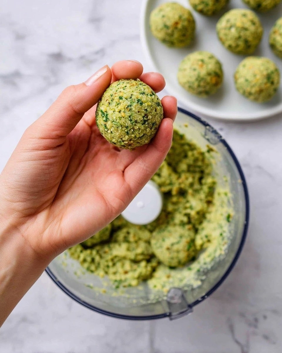 A woman's hand is holding a round falafel ball that is light green with visible bits of herbs and spices. Below the hand, there is a clear food processor bowl filled with a similar green falafel mixture smooth in texture but with small pieces throughout. In the background, on a white plate, there are several more falafel balls of the same size and color. The whole scene is set on a white marbled surface. photo taken with an iphone --ar 4:5 --v 7