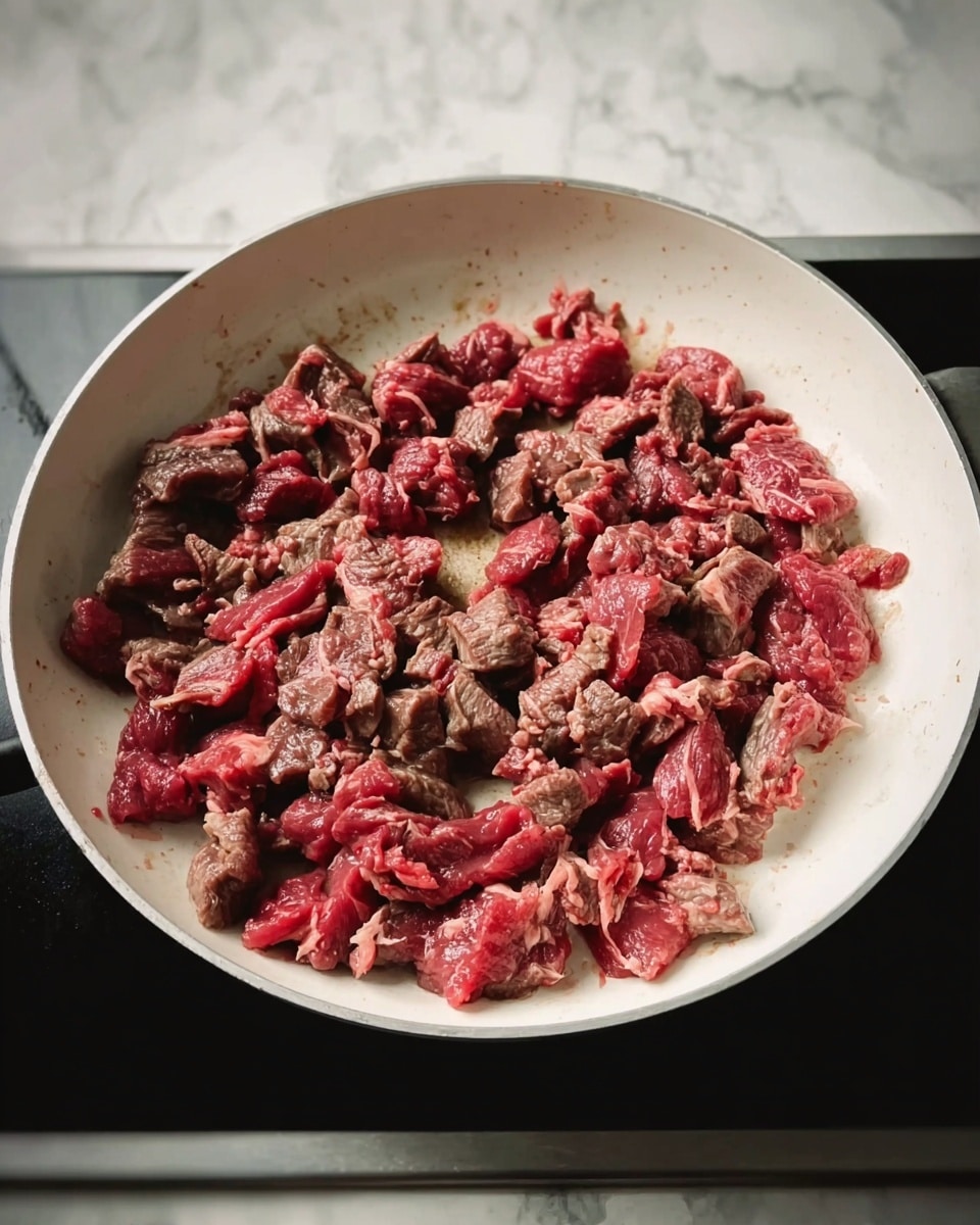 A white pan filled with small pieces of partially cooked beef, showing a mix of brown cooked meat and red raw meat. The beef pieces are scattered evenly across the bottom of the pan. The pan is placed on a black stove with a white marbled surface behind it. Photo taken with an iphone --ar 4:5 --v 7