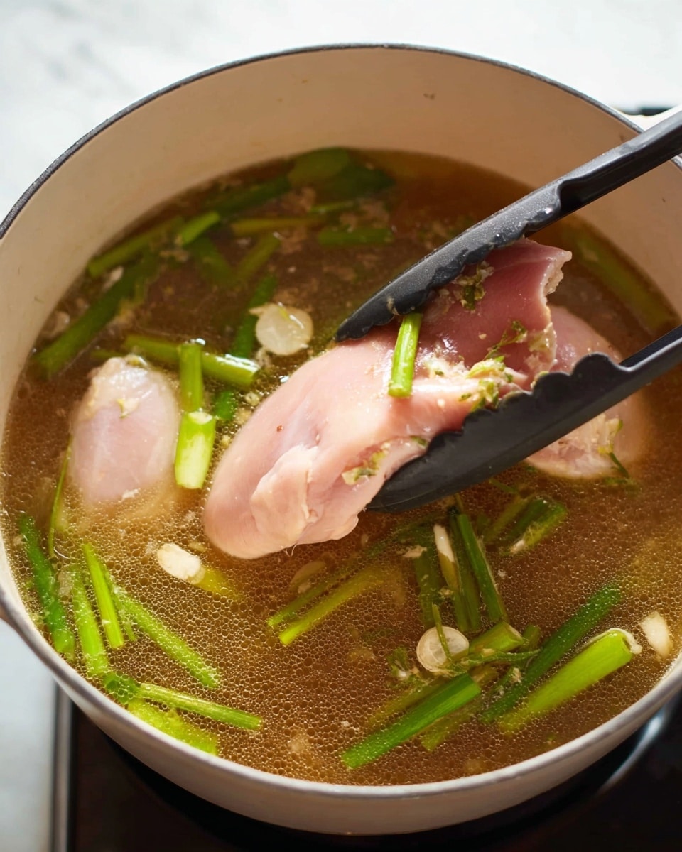 A white pot filled with clear brown broth, showing two large raw pink pieces of chicken submerged inside. The broth contains scattered bright green stalks of chopped green onions and a few small light garlic slices floating evenly around. A pair of black tongs is holding one piece of chicken above the broth near the pot edge. The pot sits on a stove over a white marbled surface. photo taken with an iphone --ar 4:5 --v 7