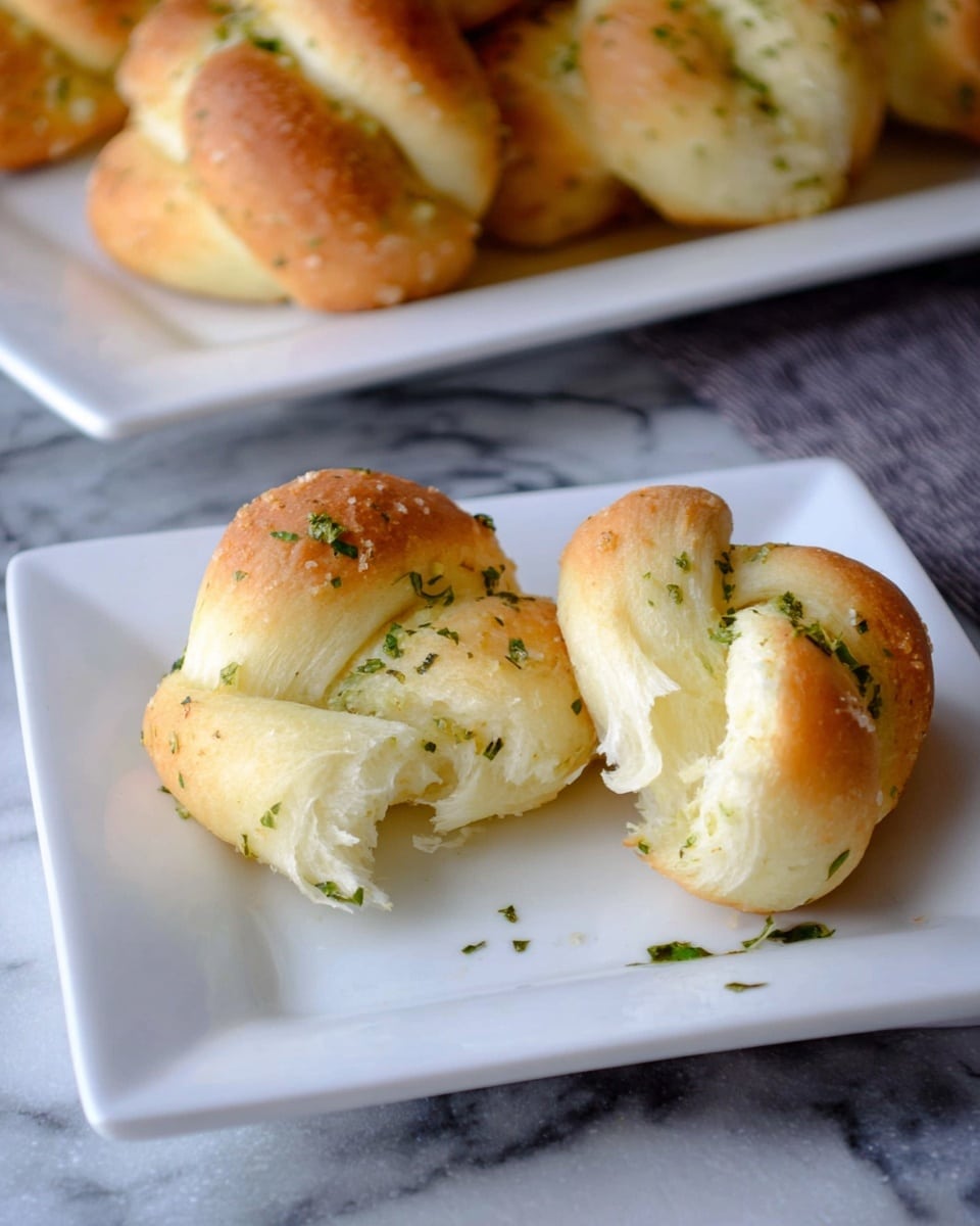 The image shows a white square plate with two soft, golden-brown garlic knots on it. One knot is whole, and the other is slightly pulled apart, exposing the fluffy white inside. Both knots have small green herbs sprinkled on top, giving a fresh look. The plate is placed on a white marbled surface, and in the background, there is a white rectangular plate holding more garlic knots, slightly out of focus. photo taken with an iphone --ar 4:5 --v 7