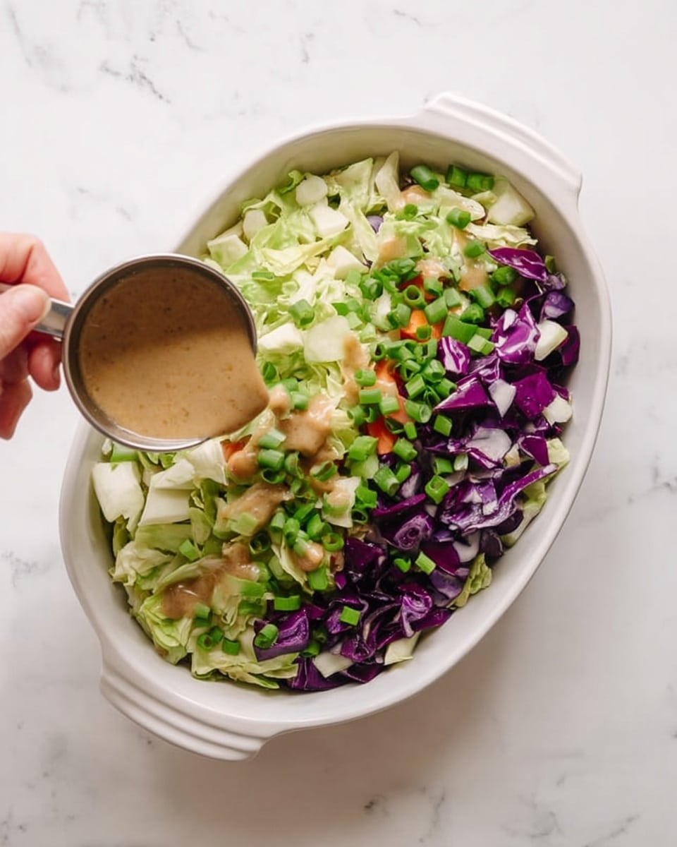 A white oval dish filled with a mix of chopped vegetables arranged in layers: a base of light green cabbage pieces, topped with chopped green onions scattered throughout, and chunks of deep purple cabbage on one side. A woman’s hand holds a small metal cup pouring a thick, light brown dressing over the vegetables. The dish sits on a white marbled surface. photo taken with an iphone --ar 4:5 --v 7