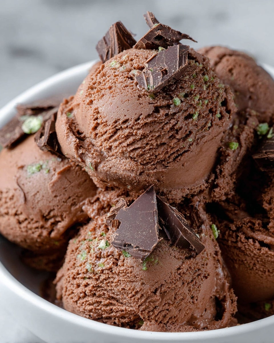 A close-up view of a silver spoon holding a scoop of creamy chocolate ice cream with small dark chocolate chunks mixed in. The ice cream has a smooth, soft texture with a light melting effect around the edges. The spoon is hovering over a white bowl filled with more chocolate ice cream, showing a dense, fluffy pile of the same dessert. The background reveals another white bowl also filled with chocolate ice cream, placed on a surface with a white marbled texture. photo taken with an iphone --ar 4:5 --v 7