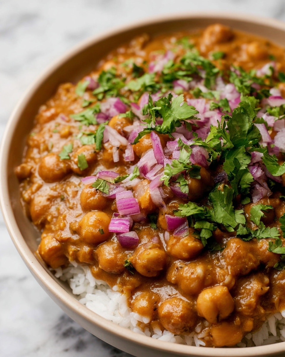 The dish shows a close-up of a white bowl filled with a creamy, brown chickpea curry layered over white rice. The curry has a thick texture with whole chickpeas visible, topped with small pieces of purple-red chopped onion and fresh green cilantro leaves scattered evenly on the top. The bowl sits on a white marbled surface, and the photo focuses on the rich textures and colors of the curry and toppings photo taken with an iphone --ar 4:5 --v 7